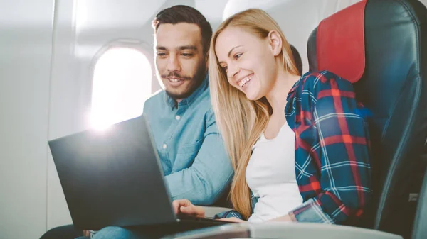 På et Board of Commercial Airplane Beautiful Young Blonde med Handsome Hispanic Male Watch Movies på en Laptop and Laugh. Sola skinner gjennom flyvinduet . – stockfoto
