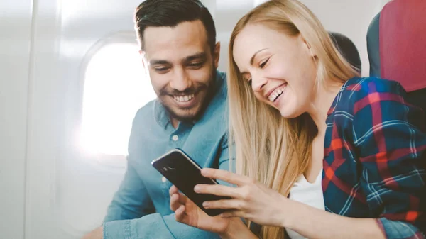 I et Board of Commercial Airplane Beautiful Young Blonde med Handsome Hispanic Male Watch Social Media på Smartphone og Le. Sola skinner gjennom flyvinduet . – stockfoto