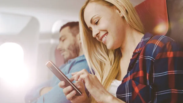 I Board of Commercial Airplane bruker vakre unge blonde Smartphone mens hennes latinamerikanske nabo sover. Sola skinner gjennom flyvinduet. . – stockfoto