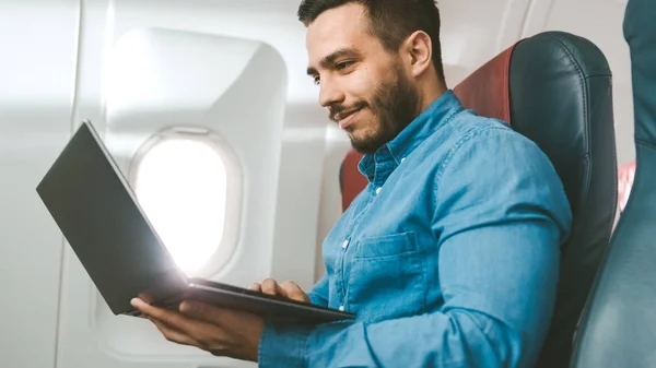 Op een bord van commerciële vliegtuig knappe Spaanse mannelijke werken op zijn Laptop. Zon schijnt door vliegtuig raam. — Stockfoto