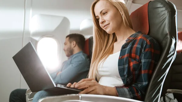 A bordo de avión comercial hermosa joven rubia trabaja en un ordenador portátil mientras su vecino hispano masculino duerme . — Foto de Stock