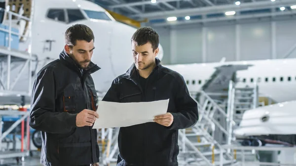 Aircraft Maintenance Worker and Engineer having Conversation. Holding Project Blueprint.