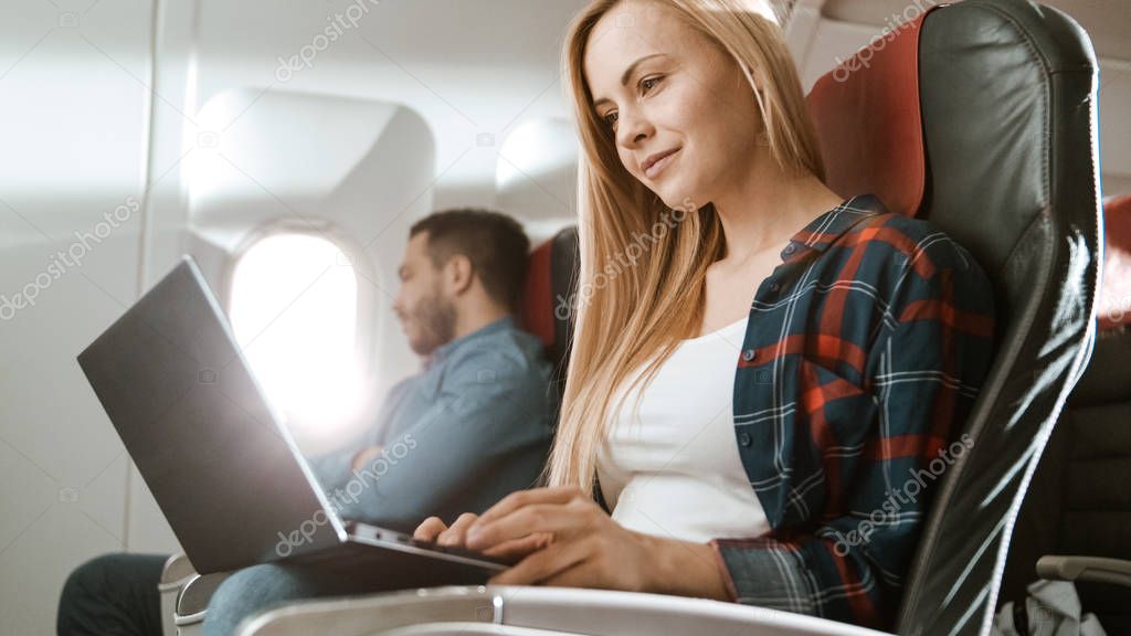 On Board of Commercial Airplane Beautiful Young Blonde Works on a Laptop while Her Hispanic Male Neighbor Sleeps.