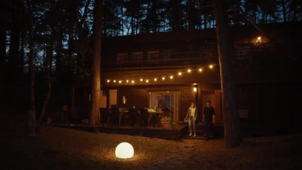 Junger Mann und Frau gehen gemeinsam von einem gemütlichen Landhaus. Haus hat warmes Licht über der Terrasse hängen. romantische Sommerabend-Atmosphäre. Ferienhaus liegt in einem Kiefernwald. — Stockvideo