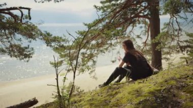 Siyah giyinmiş genç güzel kadın Bir Avrupa Çam Ormanı oturur. Doğada Sıcak Güneşli Bir Gün. O Gün Kenarlarında Dreams ve Deniz Manzara bakar.