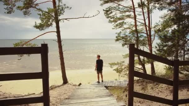 Giovane uomo è in piedi su una piattaforma rurale di legno sul bordo di una foresta di pini e guardando un bellissimo paesaggio blu del mare. Giornata calda e soleggiata con la natura. Indossa una maglietta e dei pantaloncini. . — Video Stock