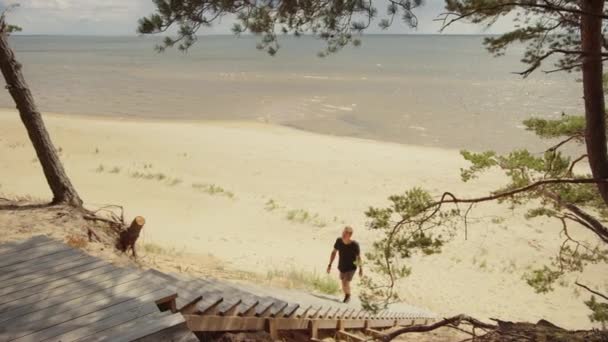 Jeune homme montant les escaliers en bois sur le bord d'une forêt de pins et sortant d'un magnifique paysage marin bleu. Journée ensoleillée chaude avec la nature. Il porte un T-shirt et un short. Il est heureux et souriant . — Video