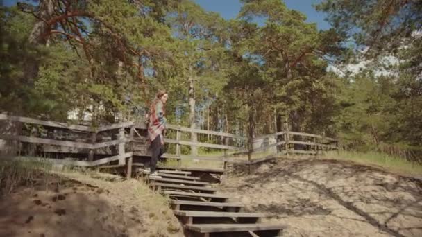 Jong mooi meisje sluit een landelijke omheining en loopt langs houten trappen aan de rand van een dennenbos. Cottage blijft op de achtergrond. Warme zonnige dag in de natuur. Ze is verpakt in een geruite Plaid. — Stockvideo