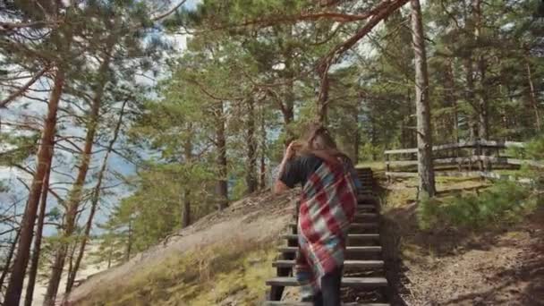 Jong mooi meisje wandelen op de houten trap aan de rand van een dennenbos en gaan van een prachtige blauwe zee landschap. Warme zonnige dag met de natuur. Ze is verpakt in een geruite Plaid. — Stockvideo