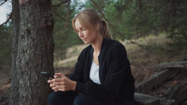 浪漫自然氛围中年轻美丽的金发女子的肖像。女孩是穿在黑色，并使用她的智能手机。她在网上浏览东西她坐在松树林里. — 图库视频影像