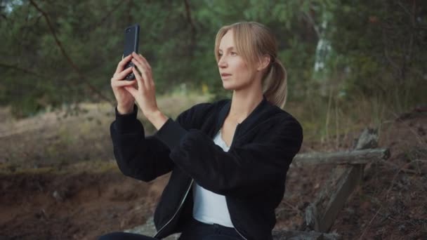 Porträtt av en ung vacker blond kvinna i en romantisk natur atmosfär. Flickan är klädd i svart och tar en bild eller en selfie på sin mobiltelefon. Hon sitter i en tallskog. — Stockvideo