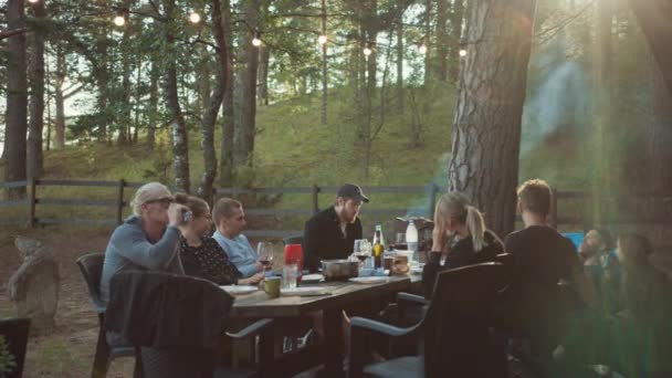 Grupo de amigos están sentados en una mesa grande en la terraza de la casa de verano en un bosque de pinos. Ambiente romántico del día de verano. Niños y niñas beben vino y otras bebidas alcohólicas ligeras. Hablan y están felices de estar vivos . — Vídeos de Stock