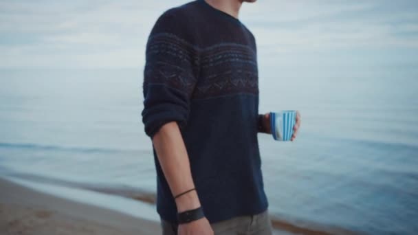 若いハンサムな男はビーチを歩きます。曇りの暖かい朝の穏やかな海。彼はティーカップから飲んでいる。彼は本物のニットセーターとショートパンツを着ている。海の近くのロマンチックな夏の時間. — ストック動画
