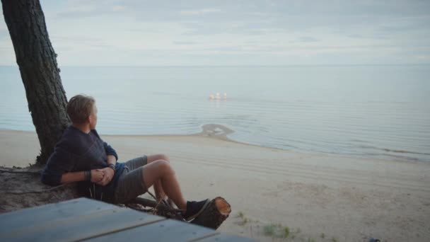 Mladý pohledný muž sedí na kraji útesu v lese. Dívá se na moře v Oblačém dni. Lidé plavou. Nosí autentický pletený svetr a šortky. Romantický letní čas. — Stock video