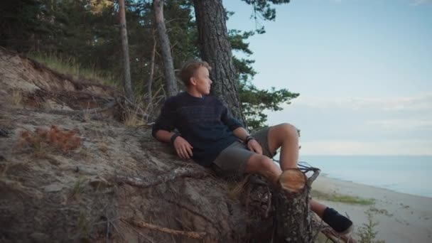 Homem bonito jovem senta-se na borda de um penhasco em uma floresta. Ele está a olhar para um mar no dia quente nublado. Ele está a usar uma camisola de malha autêntica e calções. Hora Romântica de Verão . — Vídeo de Stock