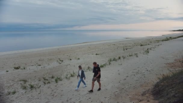 Szczęśliwa Młoda para chodzenie na opuszczonej plaży. Spokojne morze w zmierzchu wieczór Hour. Poruszają się w kierunku ognisku. Dziewczyna trzyma butelkę piwa i młody człowiek jest w szorty. Czas letni z naturą. — Wideo stockowe