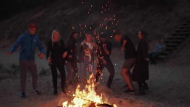 Groep vrienden dansen naast een groot kampvuur op een strand. Romantische zomeravond sfeer. Jongens en meisjes partij en drinken lichte alcoholische dranken. Ze zijn blij om te leven. — Stockvideo