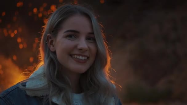 Portret van een jonge mooie blonde vrouw in een romantische Avondsfeer met een kampvuur op de achtergrond. Ze drukt een schattige glimlach uit. Vuur reflecteert op haar haar en ze draagt een barbell Earring. — Stockvideo