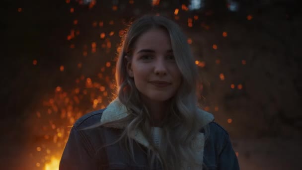 Retrato de una joven rubia hermosa en una atmósfera romántica de noche con una fogata en el fondo. Expresa una linda sonrisa. El fuego se refleja en su cabello y lleva un pendiente de la barra . — Vídeos de Stock