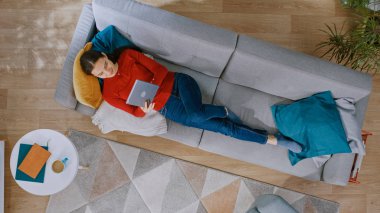Kırmızı Bluz ve Mavi Jeans Genç Kız Bir Tablet Bilgisayar kullanarak, bir Kanepede Yatıyor. O mutlu ve gülümsüyor. Halı, Masa ve Ahşap Zeminile Modern İç Mekanlı Rahat Oturma Odası. En İyi Görüntü.