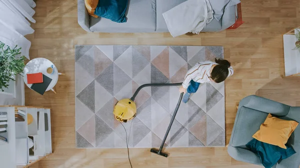 Mujer joven limpiando una acogedora sala de estar con una aspiradora. Interior moderno con alfombra, sofá, silla, mesa de centro, estante, planta y piso de madera. Arriba abajo . — Foto de Stock