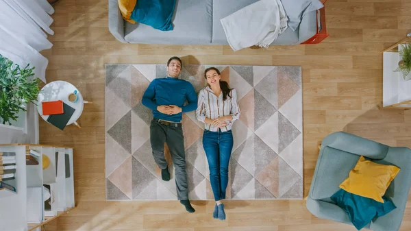 Young Couple está deitado no chão e conversando. Eles apontam os dedos para o tecto. Sala de estar acolhedora com interior moderno com tapete, cadeira, mesa, prateleira de livros, plantas e piso de madeira. Vista superior . — Fotografia de Stock