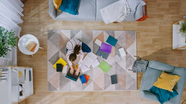 A jovem está sentada em um andar, trabalhando ou estudando em um laptop. Bebe café. Sala de estar acolhedora com interior moderno com tapete, sofá, cadeira, mesa, prateleira de livros, planta e piso de madeira. Vista superior . — Fotografia de Stock