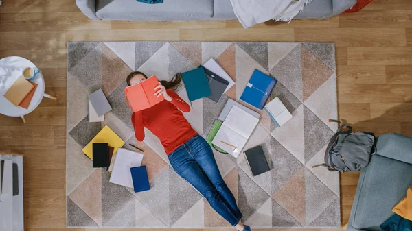 Fiatal lány a Red coat és Blue Jeans van fekve a padlón, olvasása notebook. Úgy néz ki fent, mosolyog és nevet. Cozy nappali modern belső szőnyeg, munkafüzetek és hátizsák. Felülnézet. — Stock Fotó