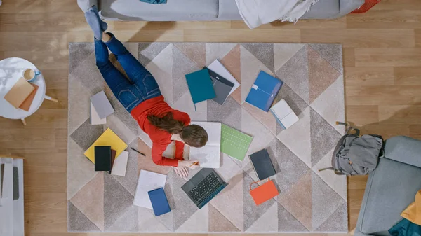 A jovem está deitada em um andar, trabalhando ou estudando em um laptop. Escreve em Notebooks. Sala de estar acolhedora com interior moderno com tapete, sofá, cadeira, mesa e piso de madeira. Vista superior . — Fotografia de Stock