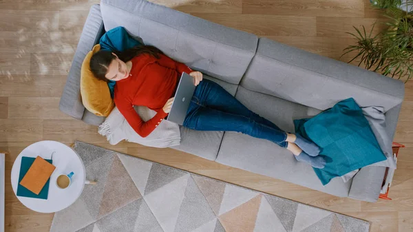 Young Girl in Red Jumper and Blue Jeans est allongé sur un canapé, travaillant sur un ordinateur portable. Regarde au-dessus, Sourires. Salon confortable avec intérieur moderne avec plantes, table basse et plancher en bois. Vue du dessus . — Photo