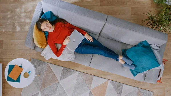 Молода дівчина в червоному перемичка і сині джинси лежачи на дивані, працюючи на ноутбуці. Виглядає вище і посміхається. Затишна вітальня з сучасним інтер'єром з килимовим покриттям, рослинами, журнальним столиком і дерев'яною підлогою. Вид зверху. — стокове фото