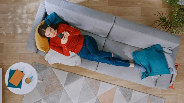 Young Girl in Red Jumper and Blue Jeans s'allonge sur un canapé à l'aide d'un téléphone intelligent. Elle est heureuse et sourit. Salon confortable avec intérieur moderne avec tapis, table basse et plancher en bois. Vue du dessus — Photo