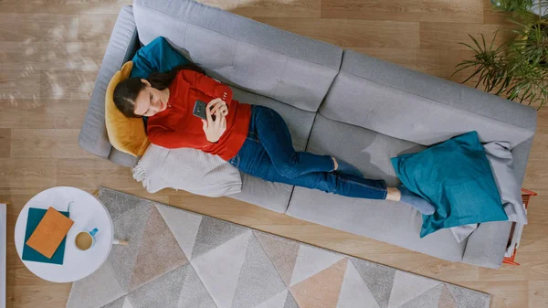 Young Girl in Red Jumper and Blue Jeans s'allonge sur un canapé à l'aide d'un téléphone intelligent. Elle est heureuse et sourit. Salon confortable avec intérieur moderne avec tapis, table basse et plancher en bois. Vue du dessus — Photo