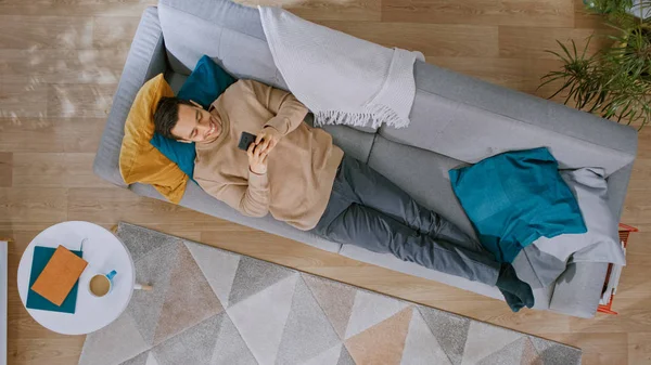 Молода людина в коричневому перемичка і сірі джинси лежачи на дивані, використовуючи смартфон. Він щасливий і посміхається. Затишна вітальня з сучасним інтер'єром з килимовим покриттям, рослинами, столиком, дерев'яною підлогою. Вид зверху. — стокове фото