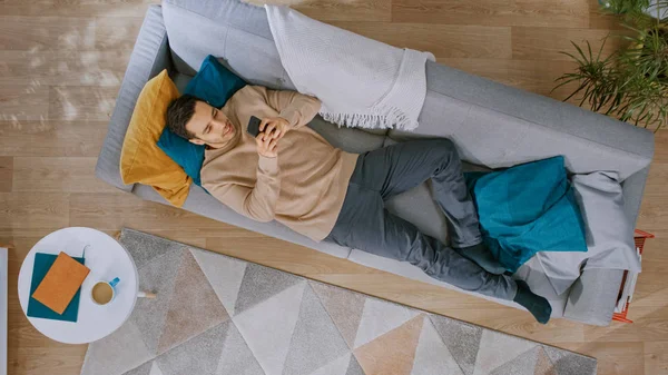 Young Man in Brown Jumper and Grey Jeans Comes and Lies Down on a Sofa, Using a Smartphone. Il est heureux et sourit. Salon confortable avec intérieur moderne avec des plantes, une table et du parquet. Vue du dessus . — Photo