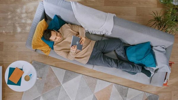 Молода людина в коричневому перемичка і сірі джинси лежачи на дивані, використовуючи планшет. Він щасливий і посміхається. Затишна вітальня з сучасним інтер'єром з рослинами, столом і дерев'яною підлогою. Вид зверху. — стокове фото