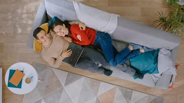 Casal bonito novo está deitado em um sofá, usando um laptop. Eles olham acima e sorriem. Sala de estar acolhedora com interior moderno com plantas, mesa e piso de madeira. Vista superior . — Fotografia de Stock