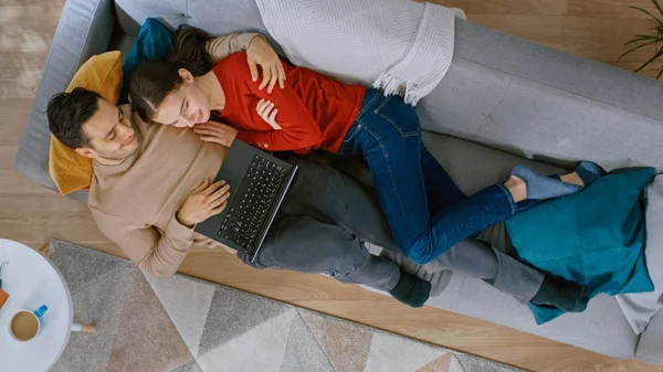 Fiatal Beautiful pár fekszik a kanapén, egy laptop. Úgy néznek ki felett és mosolyog. Hangulatos nappali modern belsővel, növényekkel, asztallal és fából készült padlóval. Felülnézet. — Stock Fotó