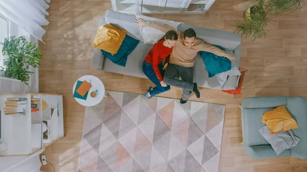 Jovem Casal Bonito Sentado em um Sofá com Travesseiros e Assistir TV. Eles estão entusiasmados e sorriem. Sala de estar acolhedora com interior moderno com plantas, mesa e piso de madeira. Vista superior . — Fotografia de Stock