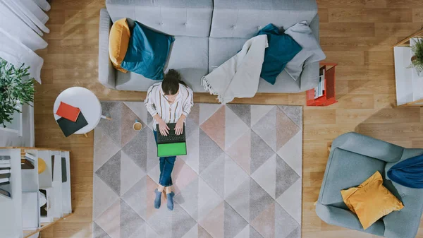 A jovem está sentada em um chão, bebendo café e usando um laptop com tela verde. Sala de estar acolhedora com interior moderno com tapete, sofá, cadeira, mesa, prateleira, planta e piso de madeira. De cima para baixo . — Fotografia de Stock