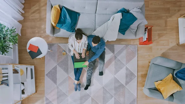 Casal jovem está sentado em um chão e usando um laptop com tela verde. Garota bebe café. Sala de estar acolhedora com interior moderno com tapete, sofá, mesa, prateleira, planta e piso de madeira. Vista superior . — Fotografia de Stock