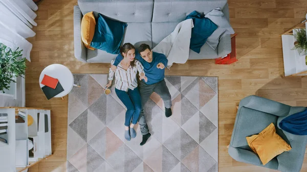 Couple est assis sur un plancher, parlant et riant. L'homme fait un câlin à la fille. Ils regardent au-dessus. Salon avec intérieur moderne avec tapis, canapé, chaise, table, étagère, plante et plancher en bois. Vue du dessus . — Photo