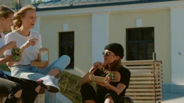 Grupo de hermosas chicas comiendo llevar comida callejera mientras se sienta en el banco en la parte Hipster con estilo de la ciudad. Tres amigas felices comiendo alimentos saludables en el parque — Vídeos de Stock