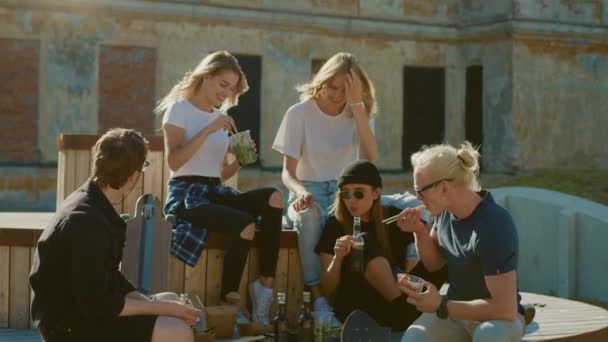 Grupp av snygga flickor och pojkar dricker hantverk drycker och äta hälsosamt ta bort gatumat på parkbänk i den svala hipster City District. Vackra unga människor som äter brunch eller lunch i parken — Stockvideo