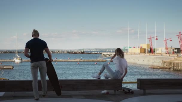 Groupe de patineurs, deux belles filles et un beau garçon monter à la jetée du port pour s'asseoir sur le banc et admirer vue sur la mer. En arrière-plan Entrepôts post-industriels et élégant quartier de Hipster — Video