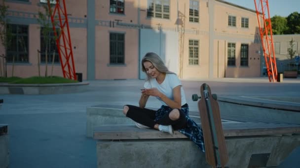 Belle patineuse blonde portant des vêtements décontractés, utilise un smartphone, partage des trucs sur les médias sociaux, prend des selfies, messages amis et s'amuser tout en étant assis sur un banc dans hippie à la mode — Video