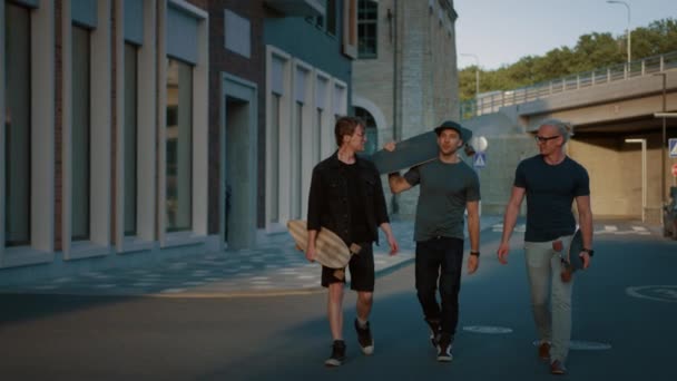 Трое крутых парней разговаривают, веселятся во время прогулок и переносят скейтборды. Стильные молодые друзья по улице. На заднем плане Модный современный район хипстеров — стоковое видео