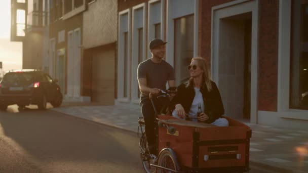 Stilig ung man rider på vuxen trehjuling med flickvän sitter i den främre lastning släpvagn. Ungt par i kärlek ha kul tillsammans ridning cykel genom fashionabla City Street — Stockvideo