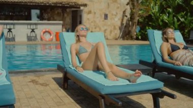 Dolly Shot Üç Güzel Genç Kadın Güneşlenme, Açık Yüzme Havuzu yakınında Şezlong larda Yatarken. Göz Alıcı ve Seyahat Hedef Tabaklanmış Başlarken Bir Mayo Muhteşem İnce Kızlar
