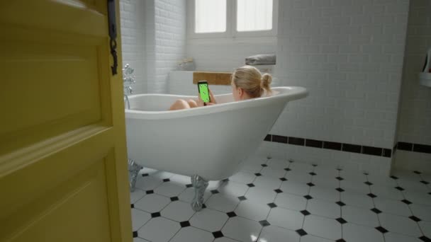 Menina loira bonita toma um banho usa verde Mock-up Screen Smartphone enquanto relaxa. Menina atraente usa Chroma Key Mobile Phone para navegar nas mídias sociais, assistir a vídeos, navegar na Internet — Vídeo de Stock